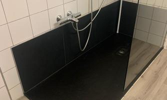 A modern shower with a black floor and tiled walls. There is a showerhead and a transparent partition panel.