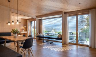 A modern living room with wooden paneling and large windows. There is a dining area and a view of the terrace.