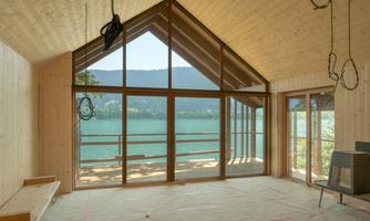 A modern designed room with large windows that offers a view of a lake and mountains. The interior is still under construction with wooden cladding and visible cables.