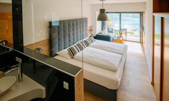 A modern hotel room with a large bed and a cozy sofa. The windows offer a view of nature and plenty of daylight.