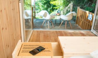 A modern furnished room with wooden-colored furniture and a terrace in the background. On a table lies a smartphone and you can see a green environment.