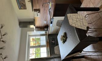 A modern kitchen with a table in the middle and chairs. The walls are bright, and there are wooden elements as well as large windows that let in plenty of daylight.