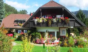 A charming house with a balcony full of flowers. The garden is colorful and well-maintained, surrounded by green trees.