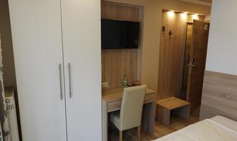 A simple room with a desk, a chair, and a wardrobe. A television hangs on the wall and the room is well-lit.