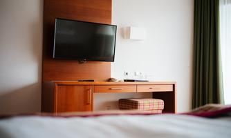 A modern bedroom with a television on the wall. A wooden piece of furniture with a padded stool is also visible.