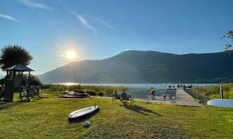 A beautiful lake with a wooden pier and gentle hills in the background. The sun is shining brightly, and there are people swimming and relaxing in the water.
