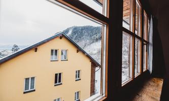 A view from a window of a yellow house and snow-covered mountains. The sky is cloudy, and the scene appears cozy and rural.