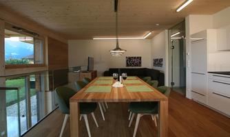 A modern dining room with a large wooden table and green chairs. The room has large windows overlooking the outside and is bright and friendly in design.