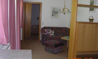 A cozy living room with a colorful sofa and a small table. The walls are light and decorated with pictures, while the windows are fitted with pink curtains.