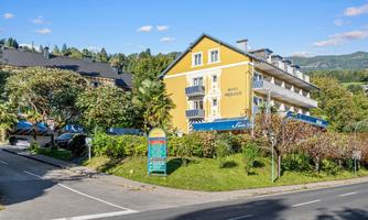 A charming hotel with a yellow façade and adjacent trees. The surroundings are green and hilly, ideal for a relaxing stay.
