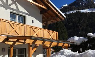 A cozy holiday home in the snow with a wooden veranda. In the background, snow-capped mountains and a blue sky can be seen.