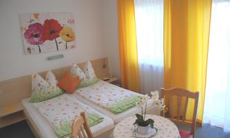 A cozy bedroom with two beds and colorful cushions. The room has large windows with yellow curtains and a small table with a flower.