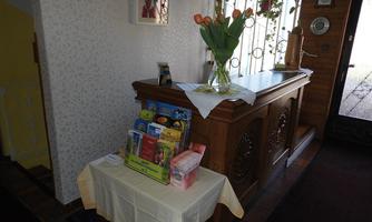 A cozy entrance area with a wooden cabinet and a tablecloth. On the table, there is a flower vase and various information sheets.