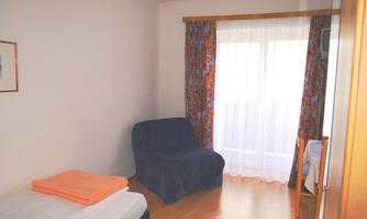 A bright room with a window and curtains. It contains a bed, a blue armchair, and a wooden chair.