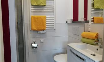 A modern bathroom with a shower, a white sink, and stylish towels in yellow and green. The walls are designed in neutral colors with red accents.