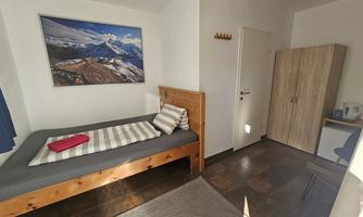 A simple bedroom with a single bed and a wooden chest of drawers. On the wall hangs a picture of the mountains, and there is a small table with a kettle.