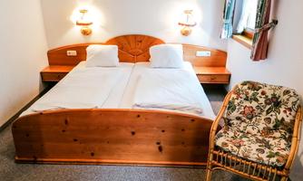 A small room with a double bed and white bedspreads. Next to the bed is an armchair with a floral design.