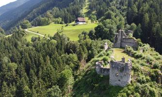 An old castle ruin on a hill, surrounded by dense forests. In the background, a house and a green meadow can be seen.