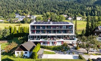 A modern building with several floors, surrounded by green landscape and trees. Guests are sitting on the terrace enjoying the view.