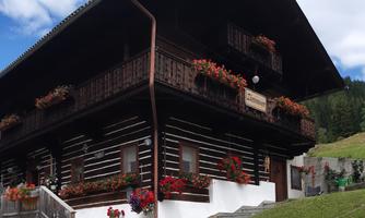A traditional wooden house with a balcony and blooming plants. The surroundings are green and hilly, ideal for a relaxing stay.
