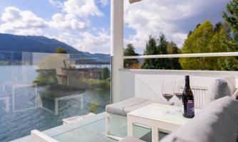 A cozy balcony with a view of a lake and surrounding mountains. On the table is a bottle of wine and two wine glasses.