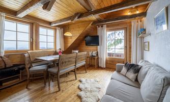 A cozy living space with wooden ceilings and large windows. A dining area and a comfortable couch invite you to relax.