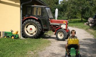 A red tractor stands next to a building, while a boy is sitting on a small green toy tractor. In the background, some trees and a green meadow can be seen.