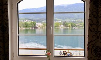 A window view of a tranquil lake with mountains in the background. A small flower stands on the windowsill.