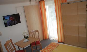 A cozy room with a double bed, a table, and two chairs. There is a television and large windows with orange curtains.
