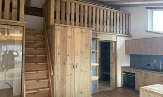 A modern room with wood paneling and a cozy loft. A wooden staircase leads to the upper floor, next to a kitchenette.