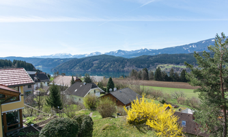 A picturesque landscape with houses, mountains, and a clear lake. The sky is blue and nature is in bloom in spring.