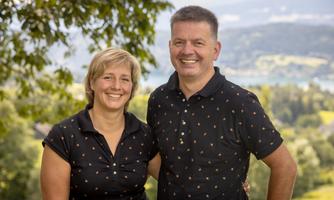 A smiling couple stands outdoors, surrounded by trees and a beautiful landscape. In the background, gentle hills and a lake are visible.