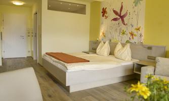 A cozy bedroom with a large bed and colorful flower motifs on the wall. The room has bright colors and an inviting atmosphere.