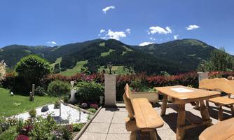 An idyllic landscape with green mountains and a blue sky. In the foreground, wooden furniture is placed in a well-kept garden.