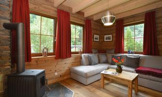 A cozy living room with a gray sofa and a wooden table. Large windows let in plenty of light, and the room features warm, rustic elements.
