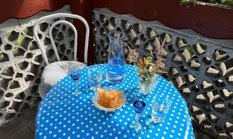 A blue tablecloth with white dots is on a small table on the terrace. On the table are a glass, a flower vase, and fruit.