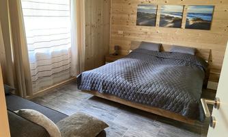 A cozy bedroom with a large double bed and gray bedding. Pictures of a landscape hang on the wall, and a window with curtains lets in daylight.