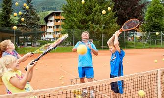 A group of children is playing tennis on a sand court. Balls are flying through the air as they try to hit them with rackets.