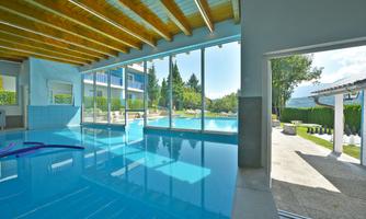 An indoor pool with large windows that provide a view of a garden with an outdoor terrace and another pool. The decor is modern and bright.