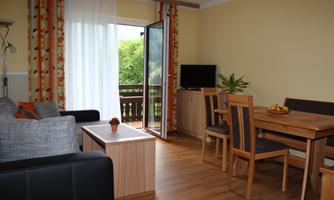 A cozy living room with a gray sofa and a wooden dining table. Large windows provide plenty of daylight and a view of greenery.