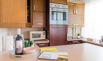 A modern kitchen with wooden cabinets and a stainless steel microwave. On the table are a bottle of wine, two cans, and some magazines.