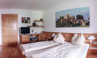 A cozy room with a large bed and wooden details. A picture of a castle hangs on the wall, and there is a desk with a television.