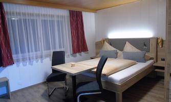 A cozy bedroom with a double bed and a table. The walls are light, and there are large windows with curtains.