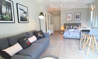 A modern living room with a gray sofa and stylish pictures on the wall. In the background, a bedroom area with a cozy bed and a table is visible.