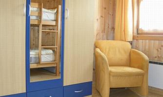 A simple room with a wardrobe and a yellow armchair. In the background, there are bunk beds visible.