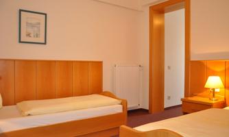 A simple room with two single beds and wooden paneling. There is a nightstand with a lamp and a window leading to an adjacent room.
