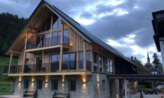 A modern wooden house with large windows and a protective canopy. The surroundings are characterized by gentle hills and wooded areas.