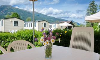 A table with a vase full of colorful flowers is in the foreground. In the background, cozy accommodations and green hills can be seen under a blue sky.