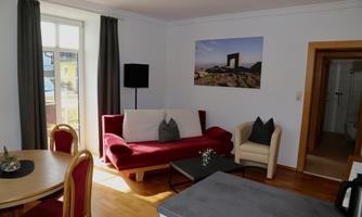A cozy living room with a red sofa and an armchair. The room has large windows and a wooden table.