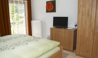A modern bedroom with a double bed and a television. The walls are bright, and there are large windows with curtains.